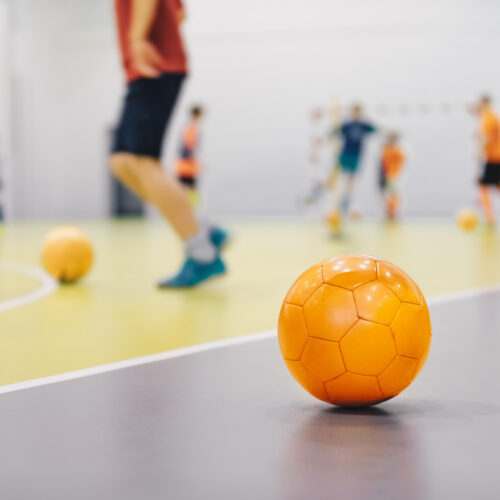 Orange Futsal Soccer Ball on Indoor Training Pitch. Young Players On Sports Practice Running Balls in Blurred Background. School Sports Class For Kids
