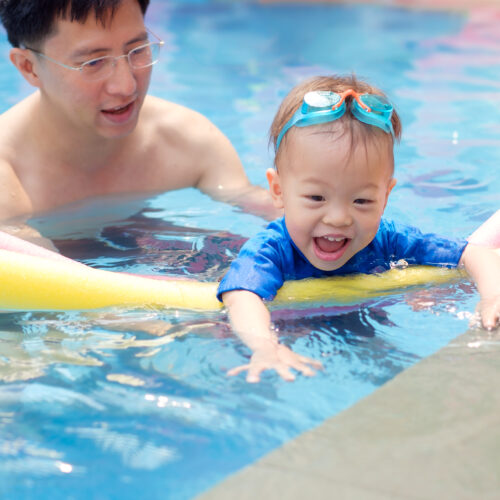 Cute little Asian 18 months / 1 year old toddler boy child wear swimming goggles learning to swim with pool noodle at outdoor pool; dad and son relaxing in swimming pool of clubhouse in summer day
