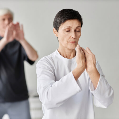 Medium shot of brunette senior Caucasian lady concentrating on qigong exercise and moving her hands, male class participants exercising in blurred background